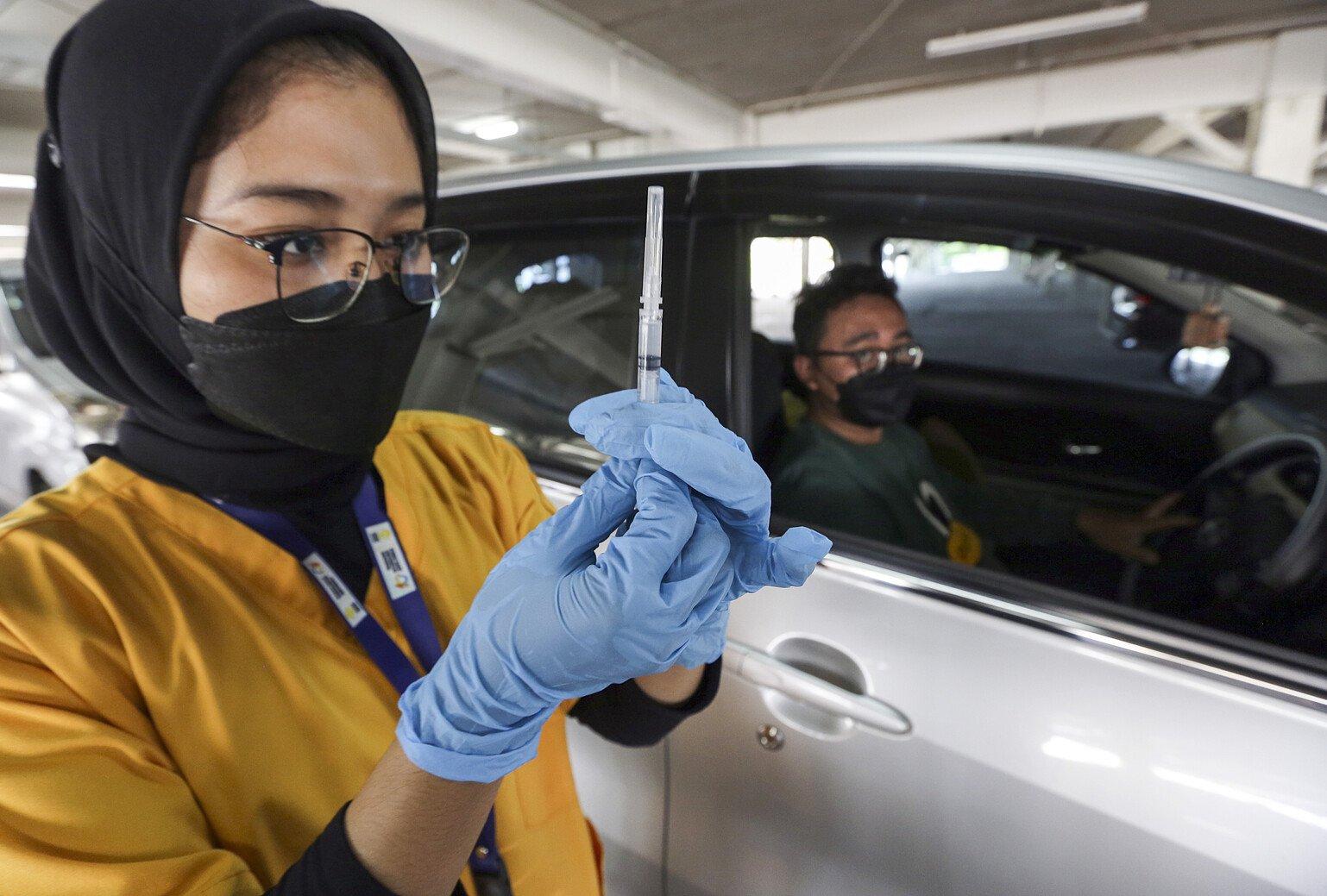 A woman with a face mask holds up a vaccine jab in the foreground while a man with a face mask sits inside a car in the background.