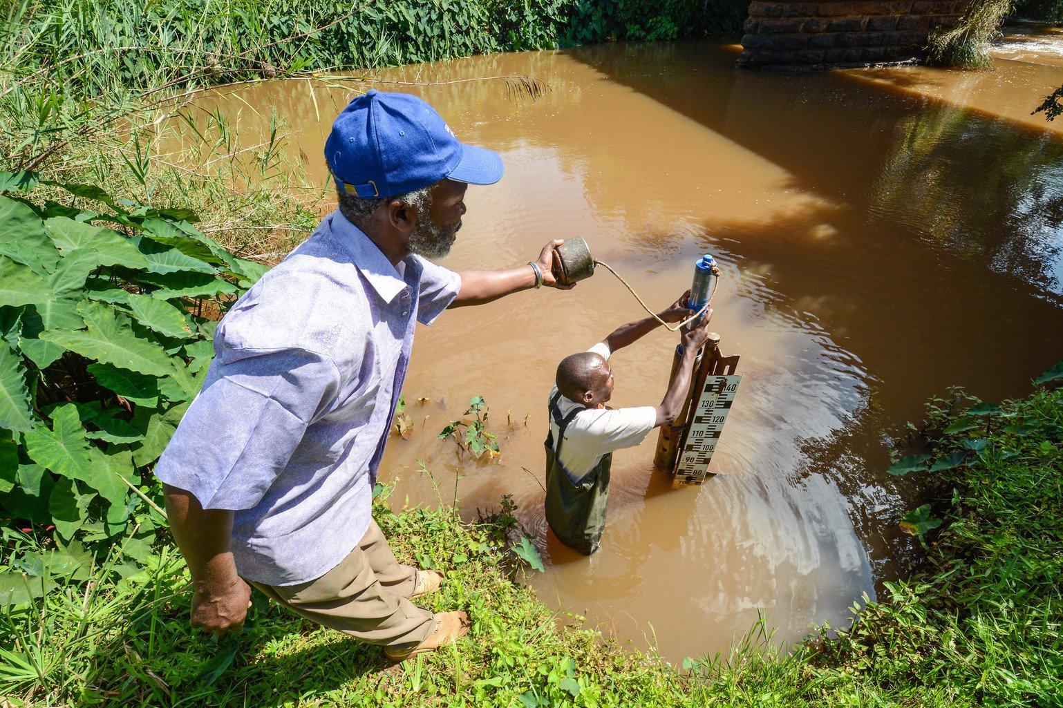 Two men are testing water from a river. One of them is in the water operating the tool, the other is standing on the bank assisting.