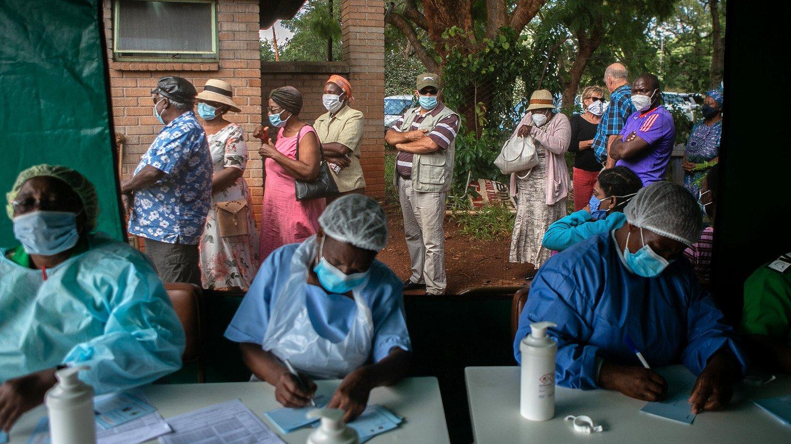 People queue for vaccination outside a tent, while three nurses write down their details on paper.