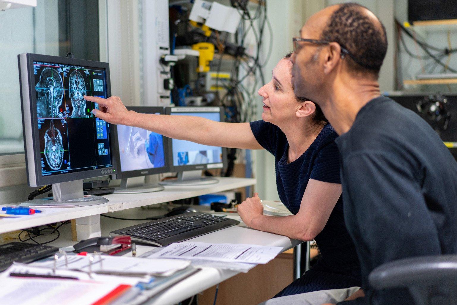 A woman talks to a patient and points at a screen showing images of a brain scan.