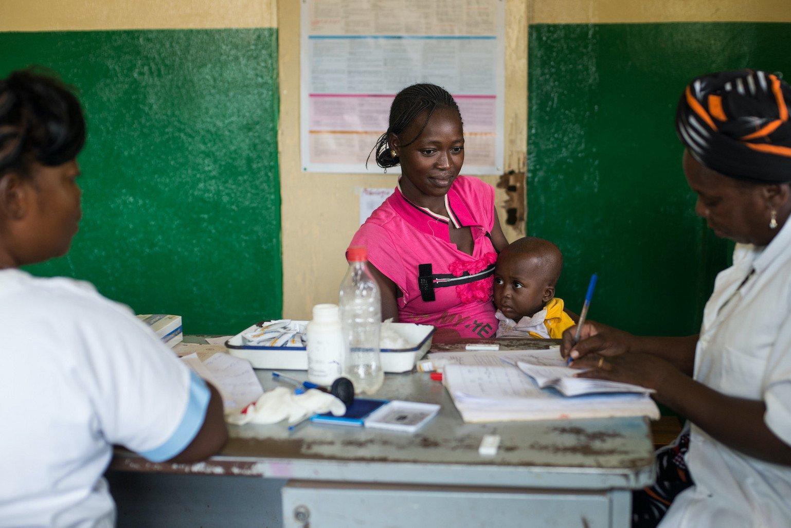 A woman wearing pink with a child on her lab sits across a desk from two other women in nurses uniform. One of the nurses is taking notes in a notebook