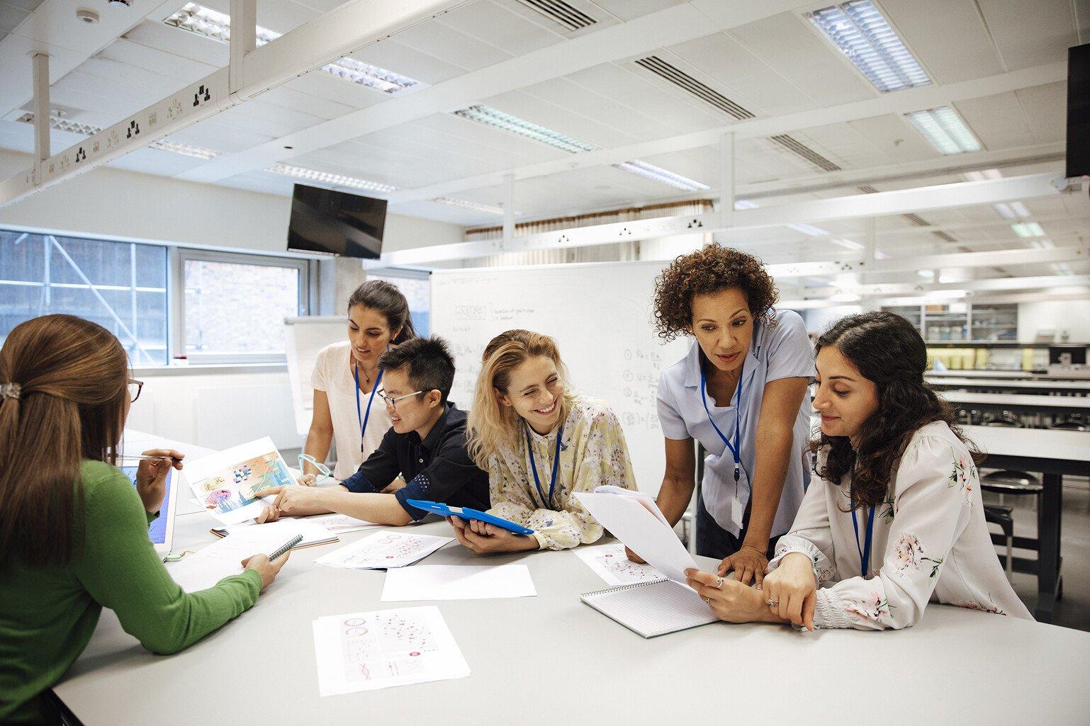 A group of researchers sit and stand around a white table, smiling and discussing some graphs.