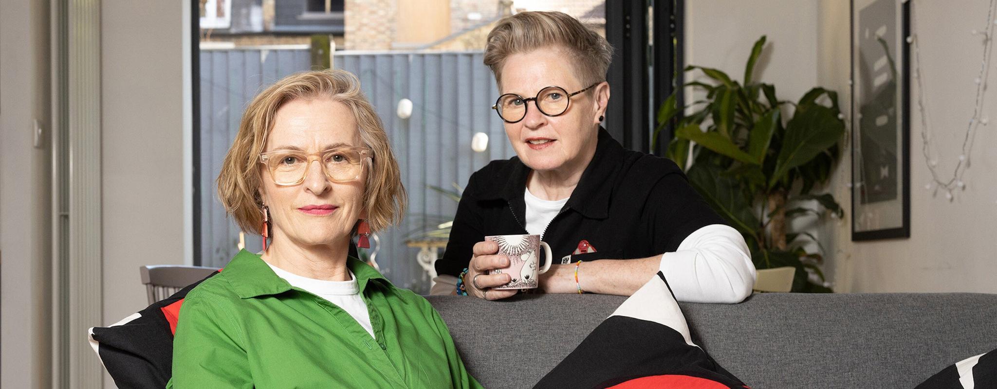 Two middle-aged women pose together in their living room. One woman in a bright green shirt sits on a grey sofa, the other sits behind her, leaning on the sofa holding a mug of tea.