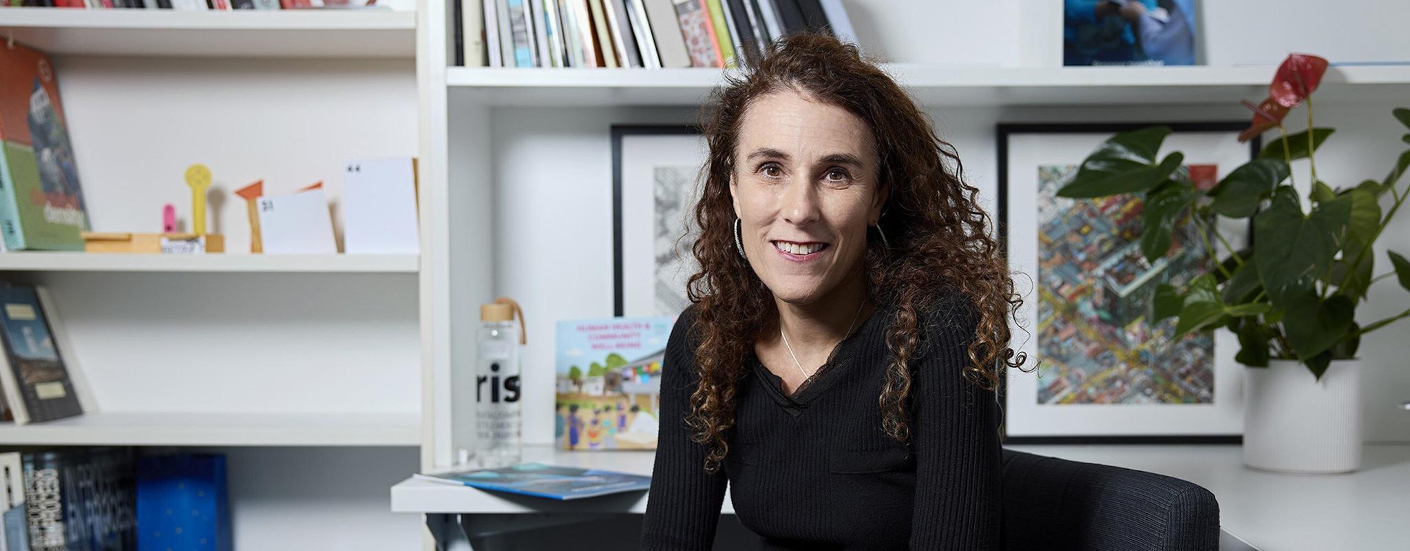 A women with dark curly hair sits in an armchair in front of a white bookcase. Her arms are crossed on her lap and she's smiling at the camera.