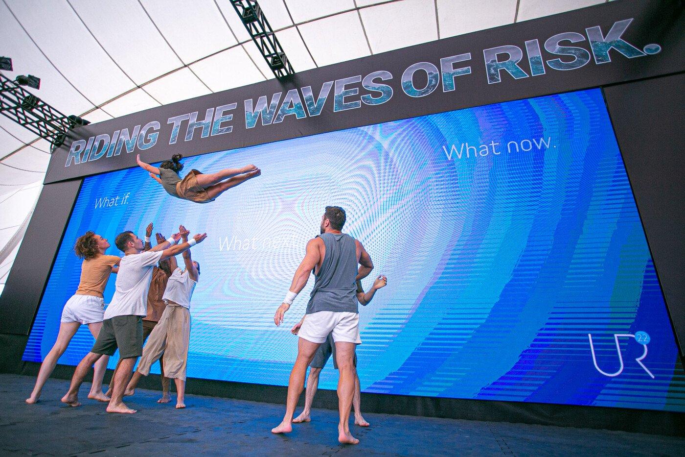 A young woman is mid air, arms and legs pointed out straight, back arched – she almost looks like a superhero. A group of four people stand below her, arms outstretched ready to catch her (presumably they have thrown her into the air as well). Several other people stand around the group, poised and ready to support if needed. It's a well rehearsed routine. The whole performance is taking place on a stage in what looks like a conference centre. A large blue backdrop reads, 'what if? what next? what now?'