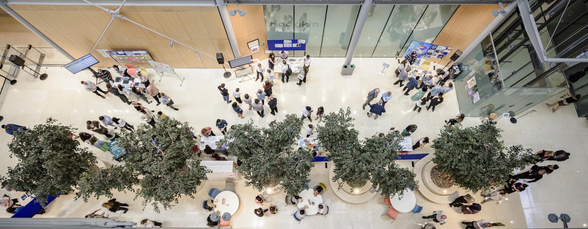 An aerial photo of the Wellcome entrance hall. Trees line the centre of a long atrium, people are gathered in small groups around information stands. It's bright and airy.