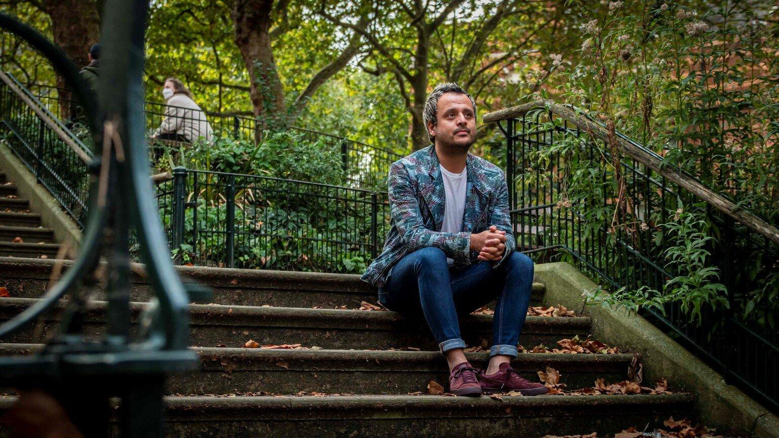 Harun Tulunay sits on some steps. He faces away to the camera, looking to the right of the frame. He wears a blue suit.