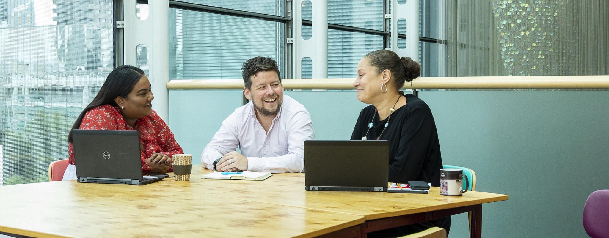 Three Wellcome staff members are sat laughing at a table with laptops. A large window overlooks the city behind them.