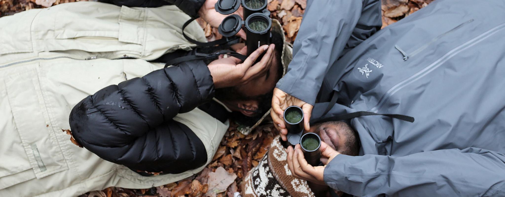 Birdwatchers lie on the ground to get a new perspective, looking upwards into the trees. 
