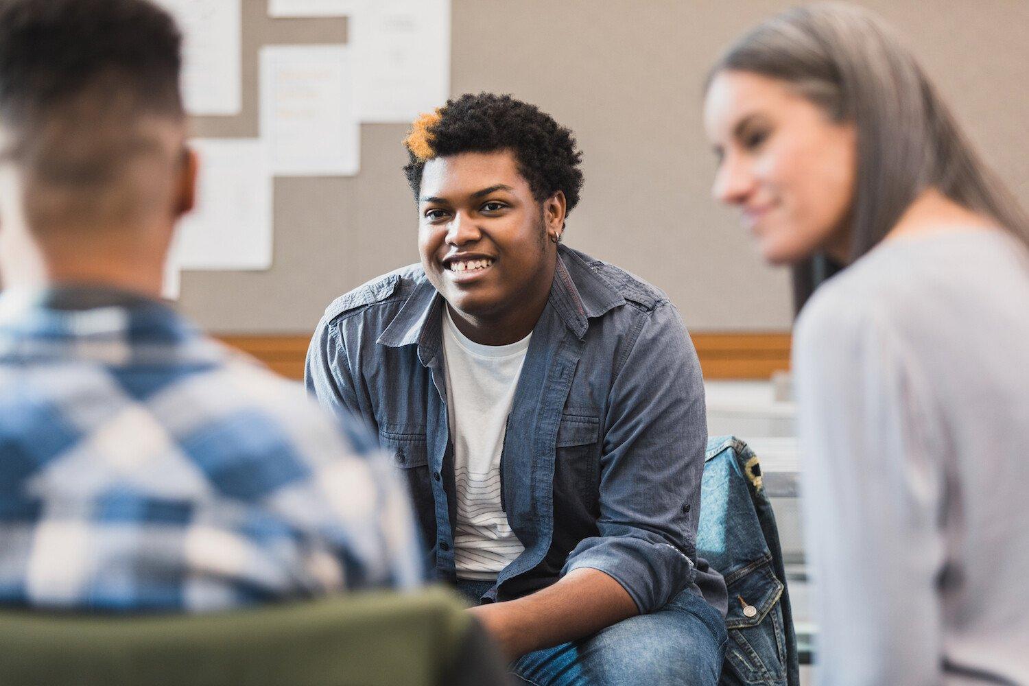 Three young people are sat in a group, smiling and listening to each other.