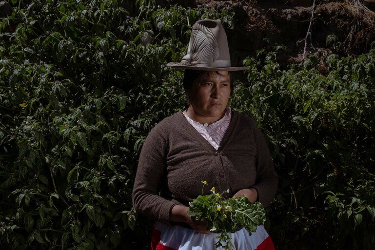 A woman in a tall hat stands holding some herbs. She wears a brown cardigan and is looking off to the right.