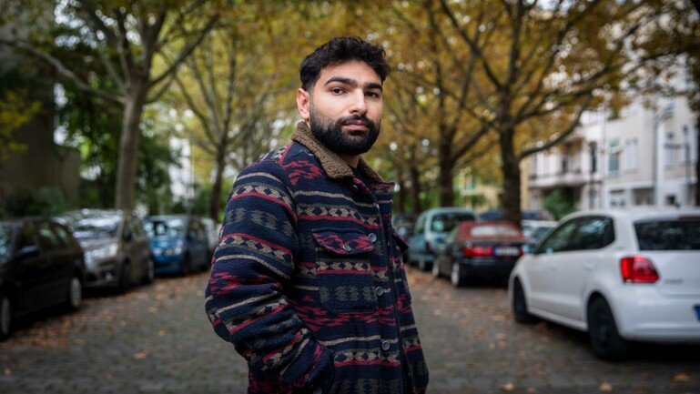 An image of a man standing on a street at centre frame. He is wearing a patterned jacket.