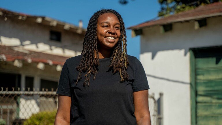 Ashley Adams, a young woman of colour stands centre frame. We see her from the waist up with her arms by her side. She wears a black t-shirt.