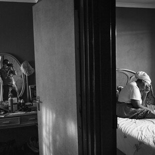 A man takes a photograph of a woman who is sitting on a bed and reading the Bible