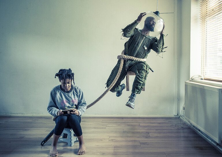 A father dressed as an astronaut floats and is tethered to his daughter who is sitting looking at her phone