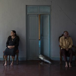 A man and a woman both sit beside an open door, with a big fish on the ground between them. 