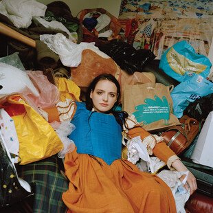 A woman sits on a bed crowded with clothes and bags.