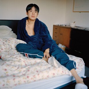 A boy lounges on a bed, a collage in progress on the desk behind him. 